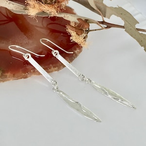 May include: A pair of silver dangle earrings with a long, thin, textured bar and a smaller, textured, leaf-shaped pendant.