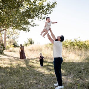 May include: A family enjoys a sunny day outdoors. A man throws a baby in the air, while another child and a woman walk in the background. The scene is set in a grassy field with trees.