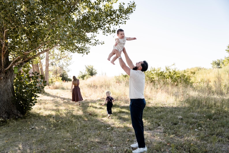 May include: A family enjoys a sunny day outdoors. A man throws a baby in the air, while another child and a woman walk in the background. The scene is set in a grassy field with trees.