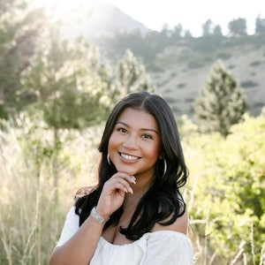 May include: A smiling person with long, dark hair and silver hoop earrings poses outdoors. They wear a white off-the-shoulder top and a silver bracelet. The background features tall grass, trees, and a mountain range, with sunlight shining through.