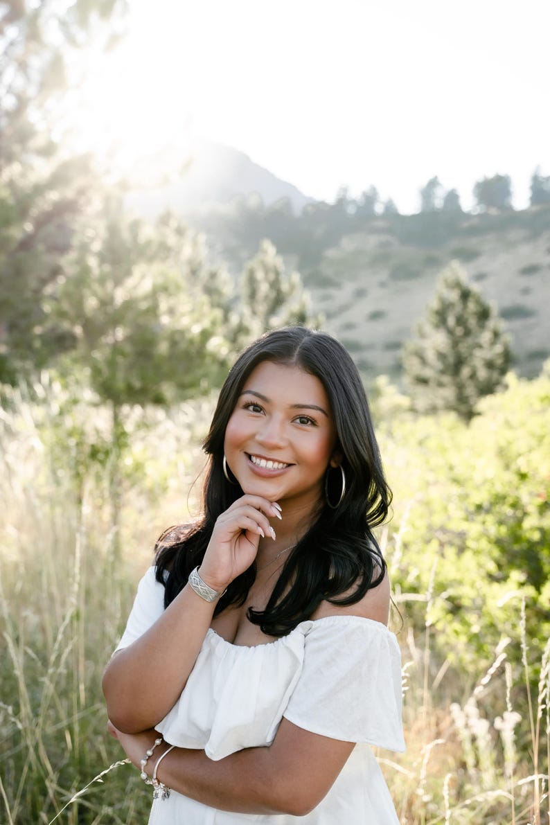 May include: A smiling person with long, dark hair and silver hoop earrings poses outdoors. They wear a white off-the-shoulder top and a silver bracelet. The background features tall grass, trees, and a mountain range, with sunlight shining through.