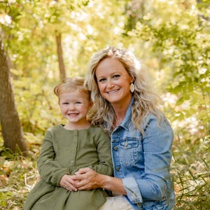 May include: A smiling woman and young girl pose outdoors in a forest setting. The woman wears a denim jacket and the girl wears a green dress. The background features lush green and yellow foliage, with a tree trunk visible on the left.