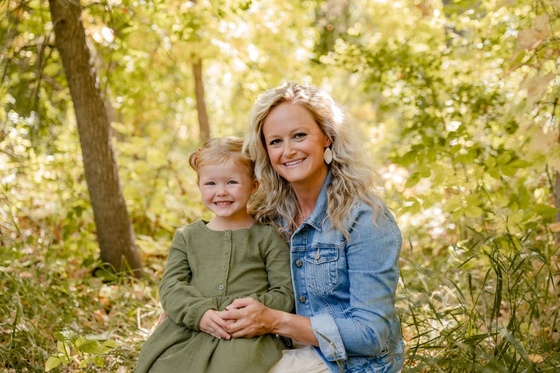 May include: A smiling woman and young girl pose outdoors in a forest setting. The woman wears a denim jacket and the girl wears a green dress. The background features lush green and yellow foliage, with a tree trunk visible on the left.