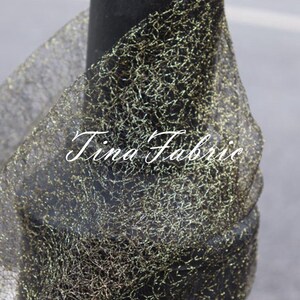 Puede incluir: Tela de malla negra con purpurina dorada tejida en todo. La tela est&aacute; drapeada sobre una superficie negra. El texto "Tina Fabric" es visible en la imagen.