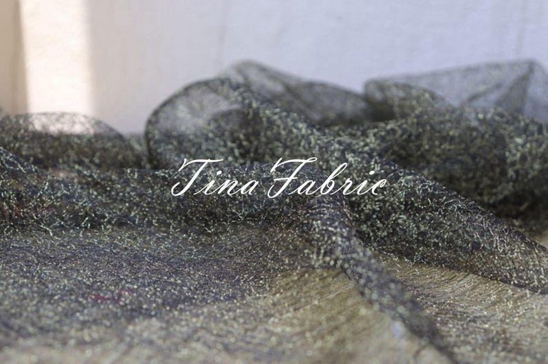 Puede incluir: Tela negra transparente con purpurina dorada tejida. La tela es texturizada y tiene un brillo sutil. El texto "Tina Fabrie" es visible en la imagen.