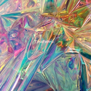 Pode incluir: Close-up de um tecido holográfico iridescente com um brilho arco-íris. O tecido está enrugado e dobrado, criando uma superfície texturizada. O texto "TinaFabric" é visível no centro inferior da imagem.