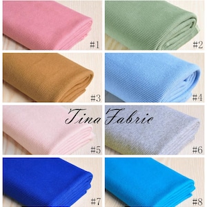 May include: Eight different colours of ribbed knit fabric, folded and displayed on a wooden surface. The colours are pink, green, brown, light blue, light pink, grey, blue, and turquoise.