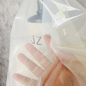 May include: Clear plastic sheet with a slight sheen. The sheet is draped over a form and a hand is holding the sheet up.