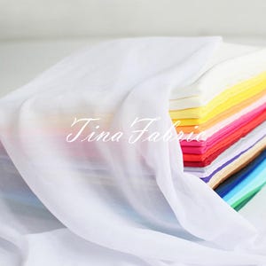Pode incluir: Uma pilha de quadrados de tecido coloridos, dobrados e sobrepostos, com um tecido branco transparente drapeado por cima. Os quadrados de tecido são em vários tons de rosa, vermelho, laranja, amarelo, verde, azul e roxo.