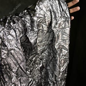 Op de afbeelding: Zilverkleurige metallic stof met een getextureerde, gekreukelde uitstraling. De stof wordt omhoog gehouden, waardoor het gekreukelde oppervlak en de manier waarop het licht vangt, zichtbaar worden. De stof heeft een donkere achtergrond.