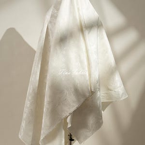 Puede incluir: Tela blanquecina con un sutil estampado floral, drapeada y exhibida sobre un soporte. La tela tiene un brillo suave y está texturizada. El texto "Tina Fabric" es visible en la tela.