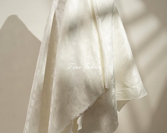 May include: Off-white fabric with a subtle floral pattern, draped and displayed on a stand. The fabric has a soft sheen and is textured. The text "Tina Fabric" is visible on the fabric.