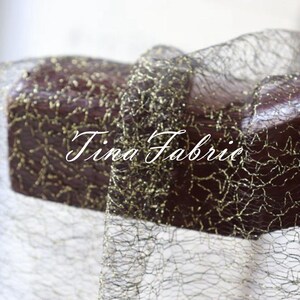 Puede incluir: Tela de malla negra con detalles de purpurina dorada. La tela est&aacute; drapeada sobre una superficie de madera. El texto "Tina Fabric" es visible en la imagen.