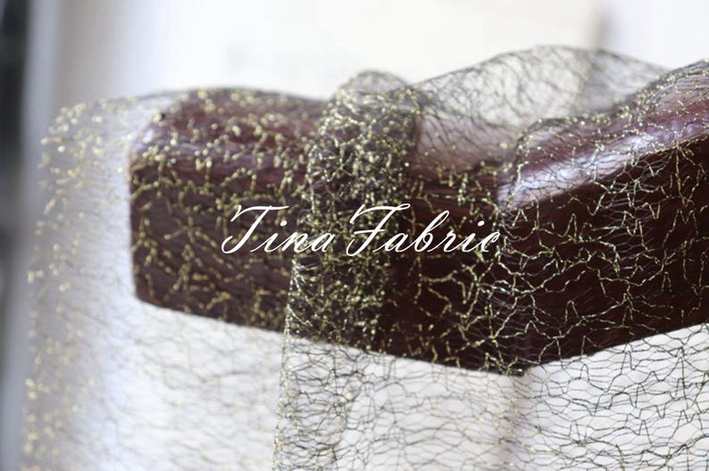 Puede incluir: Tela de malla negra con detalles de purpurina dorada. La tela est&aacute; drapeada sobre una superficie de madera. El texto "Tina Fabric" es visible en la imagen.