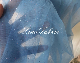 May include: Light blue tulle fabric with iridescent glitter. The fabric is sheer and has a delicate texture. The text "Tina Fabric" is visible in the lower center of the image.