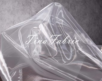 May include: White sheer fabric with a delicate, crinkled texture. The fabric is draped and folded, creating soft, flowing lines. The text "Tina Fabrie" is printed in white on the fabric.