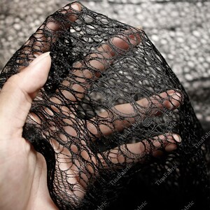 May include: Close-up of black open-weave fabric. The fabric has a net-like appearance with large, irregular holes. The material is held up, allowing light to pass through, highlighting the texture and pattern. The fabric appears to be soft and flexible.