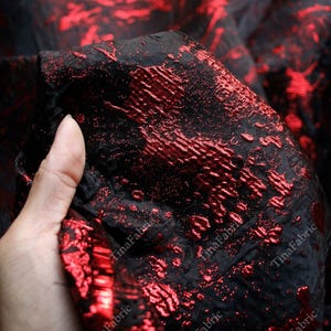 Pode incluir: Close-up de um tecido preto com um padrão texturizado e em relevo em vermelho vibrante. O tecido tem uma aparência brilhante e metálica, com o design vermelho contrastando com o fundo preto. O tecido é provavelmente usado para roupas ou decoração de casa.
