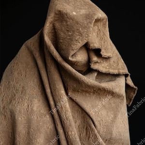 Peut inclure: Un tissu drapé et texturé de couleur marron chaud. Le tissu est plié et drapé, créant une forme de capuche. La surface présente un motif subtil en relief. Le fond est noir uni.