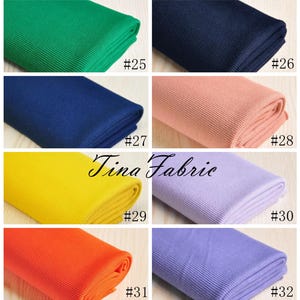 May include: A collection of ten fabric swatches in various colours, including green, blue, pink, yellow, orange, and purple. Each swatch is folded and labelled with a number from 25 to 32. The fabric appears to be ribbed.