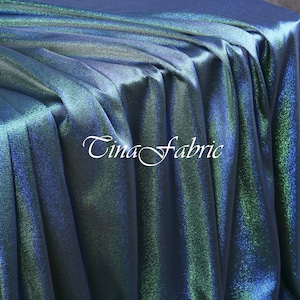 May include: A draped fabric with a shimmering, iridescent effect. The fabric transitions from deep blue to teal and green, creating a dynamic visual texture. The text "Tina fabric" is displayed in a cursive font.