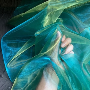 May include: Close-up of sheer fabric in shades of teal, turquoise, and chartreuse. The fabric is layered and translucent, with light passing through it. The material appears to be organza or a similar lightweight textile.