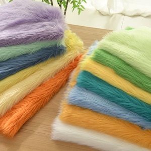 May include: A selection of colourful faux fur fabric in shades of purple, green, blue, yellow, orange, and white. The plush material has long, soft fibres, suitable for crafts or decor.