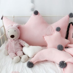 May include: A set of pink star-shaped and crescent-shaped pillows with gray pom-poms. The pillows are arranged on a white bed with a stuffed animal.