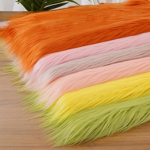 Puede incluir: Primer plano de tela de piel sintética colorida en tonos naranja, rosa, gris, amarillo y verde. Las fibras largas y suaves están dispuestas en capas, creando una exhibición texturizada y vibrante. La piel se utiliza probablemente para manualidades o disfraces.