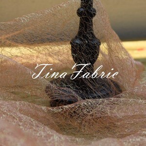 Puede incluir: Primer plano de una tela fina y transparente de color marr&oacute;n claro con un candelero negro adornado en el centro. La tela tiene una textura fina en forma de red y el candelero tiene un dise&ntilde;o decorativo e intrincado. El texto "Tina Fabric" es visible en la imagen.