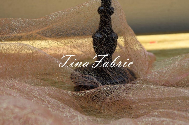 Puede incluir: Primer plano de una tela fina y transparente de color marr&oacute;n claro con un candelero negro adornado en el centro. La tela tiene una textura fina en forma de red y el candelero tiene un dise&ntilde;o decorativo e intrincado. El texto "Tina Fabric" es visible en la imagen.