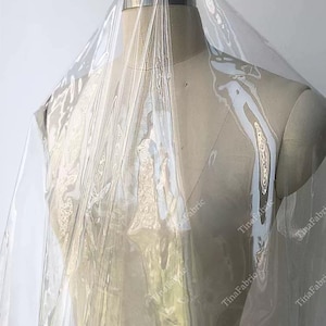 May include: A clear, transparent vinyl fabric draped over a dress form. The fabric is shiny and reflects light, creating a rippled effect. The dress form is a neutral beige color, and the background is plain.