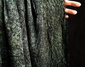 May include: Dark green fabric with a textured, crinkled appearance. The fabric has a mottled pattern with black accents, creating a marbled effect. The edge of the fabric is fringed. The fabric is held up against a dark background.