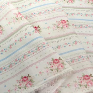 May include: Off-white fabric featuring a repeating pattern of pink roses, blue and green stripes, and pink lace trim. The fabric is folded, showing the frayed edges. The floral design gives a vintage, romantic feel.