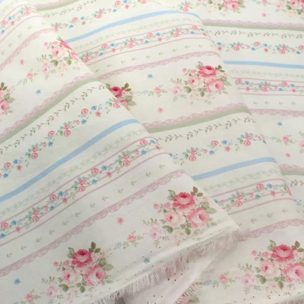 May include: Off-white fabric with a repeating pattern of pink roses, blue and green stripes, and pink lace trim. The fabric is folded, showing the frayed edges. The floral design gives a vintage, romantic feel.