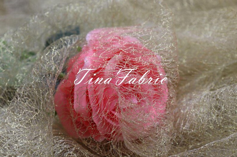 Puede incluir: Una rosa rosa envuelta en una tela de tul dorada con purpurina. La tela tiene una textura delicada y de red. El texto "Tina Fabric" es visible en la rosa.