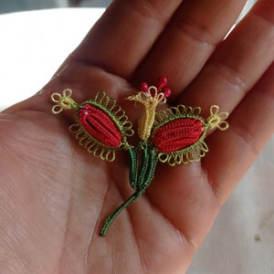 May include: A delicate crocheted flower with red, yellow, and green thread. The flower has two petals and a stem.