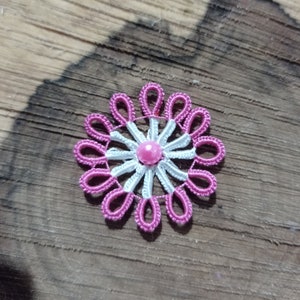 May include: A handmade, floral-shaped embellishment with a pink and white design. The flower has a central pink bead, surrounded by white and pink looped petals. The craft item is set against a wooden background.