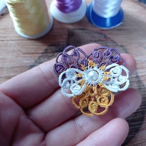 May include: A heart-shaped crocheted flower made with purple, white and yellow thread. The centre of the flower has a pearl bead.