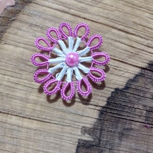 May include: A handmade, pink and white floral brooch. The brooch features a central pink pearl surrounded by white and pink thread petals. The brooch is set against a wooden background.