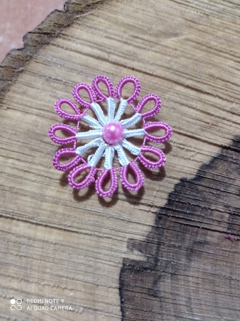 May include: A handmade, pink and white floral brooch. The brooch features a central pink pearl surrounded by white and pink thread petals. The brooch is set against a wooden background.