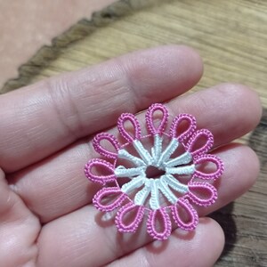 May include: A handmade, delicate, floral-shaped lace doily. The doily features a white center with pink petal-like loops around the edge. The lace is held in a hand, showcasing the intricate detail of the craft.