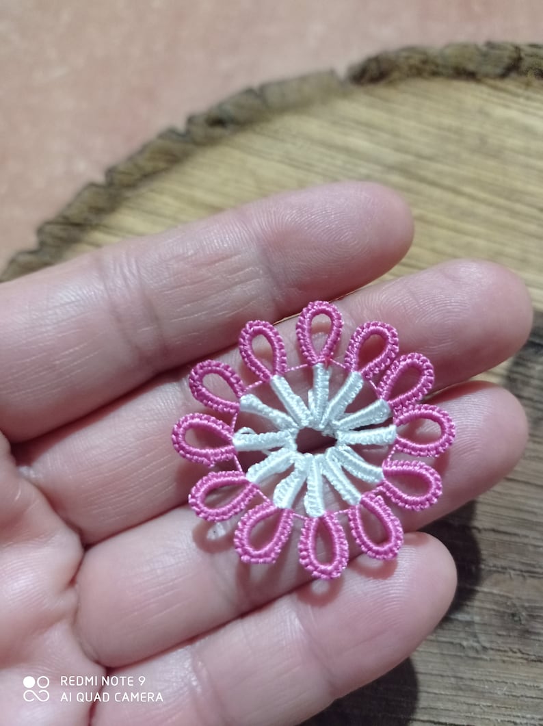 May include: A handmade, delicate, floral-shaped lace doily. The doily features a white center with pink petal-like loops around the edge. The lace is held in a hand, showcasing the intricate detail of the craft.