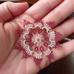 May include: A handmade, delicate lace doily in a snowflake design. The doily features a symmetrical pattern with alternating burgundy and off-white sections. The intricate lacework is displayed against a neutral background.