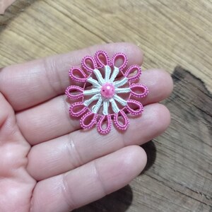 May include: A handmade, pink and white floral applique with a central pink bead. The delicate, lace-like design features a series of looped petals around a central starburst pattern. The applique is likely for embellishing clothing or accessories.