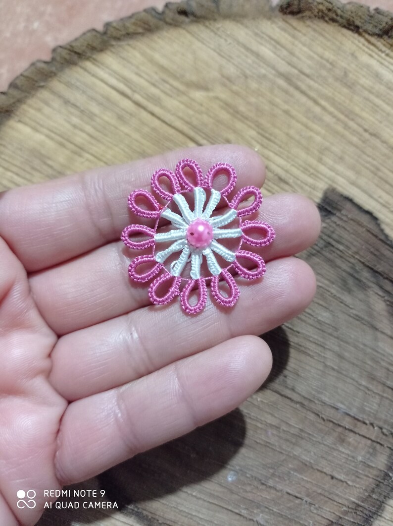 May include: A handmade, pink and white floral applique with a central pink bead. The delicate, lace-like design features a series of looped petals around a central starburst pattern. The applique is likely for embellishing clothing or accessories.