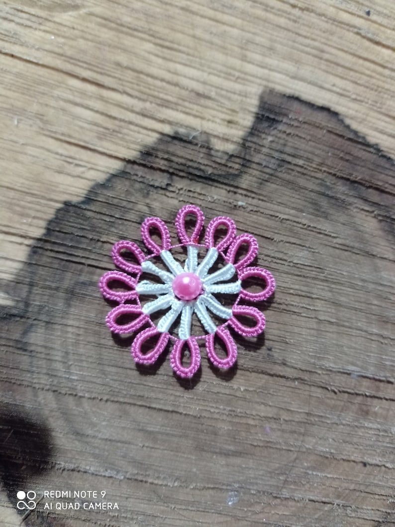May include: A handmade, floral-shaped embellishment with a pink and white design. The flower has a central pink bead, surrounded by white and pink looped petals. The craft item is set against a wooden background.