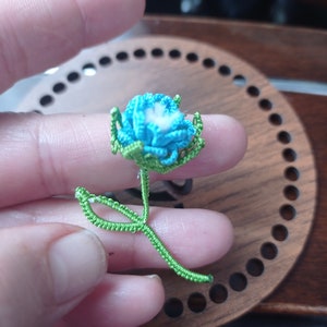 May include: A small crocheted flower with a blue center and green leaves. The flower is held in a hand.