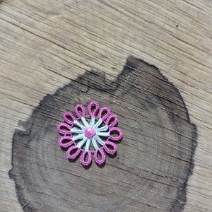 May include: A handmade, pink and white lace flower with a central pink bead. The flower is set against a natural wood background, showcasing the intricate details of the lace design. The flower's petals are a vibrant pink, and the center is a lighter pink.