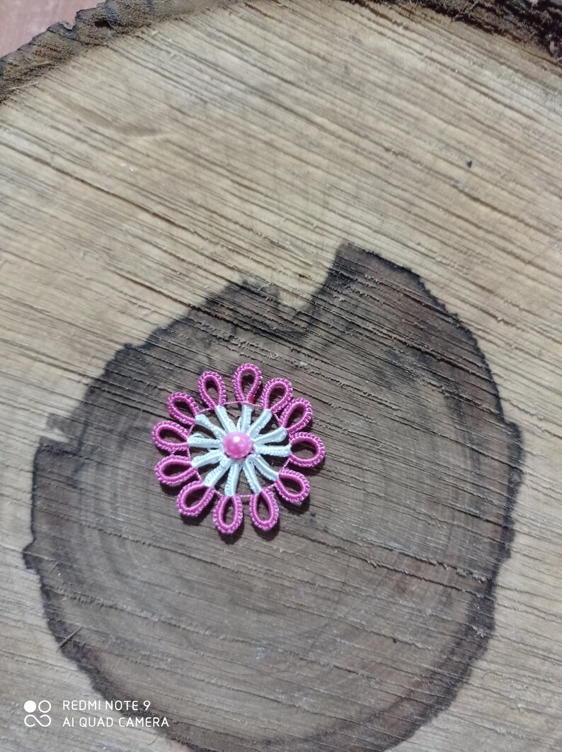 May include: A handmade, pink and white lace flower with a central pink bead. The flower is set against a natural wood background, showcasing the intricate details of the lace design. The flower's petals are a vibrant pink, and the center is a lighter pink.